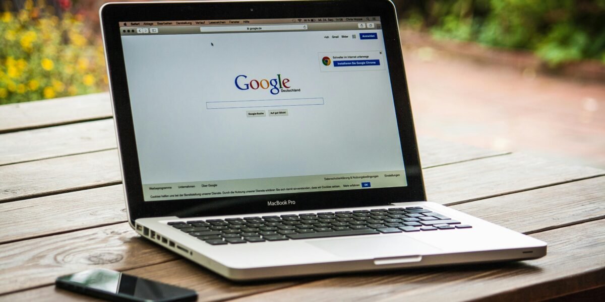 A MacBook Pro displaying Google Search on a wooden table outdoors, next to a smartphone.